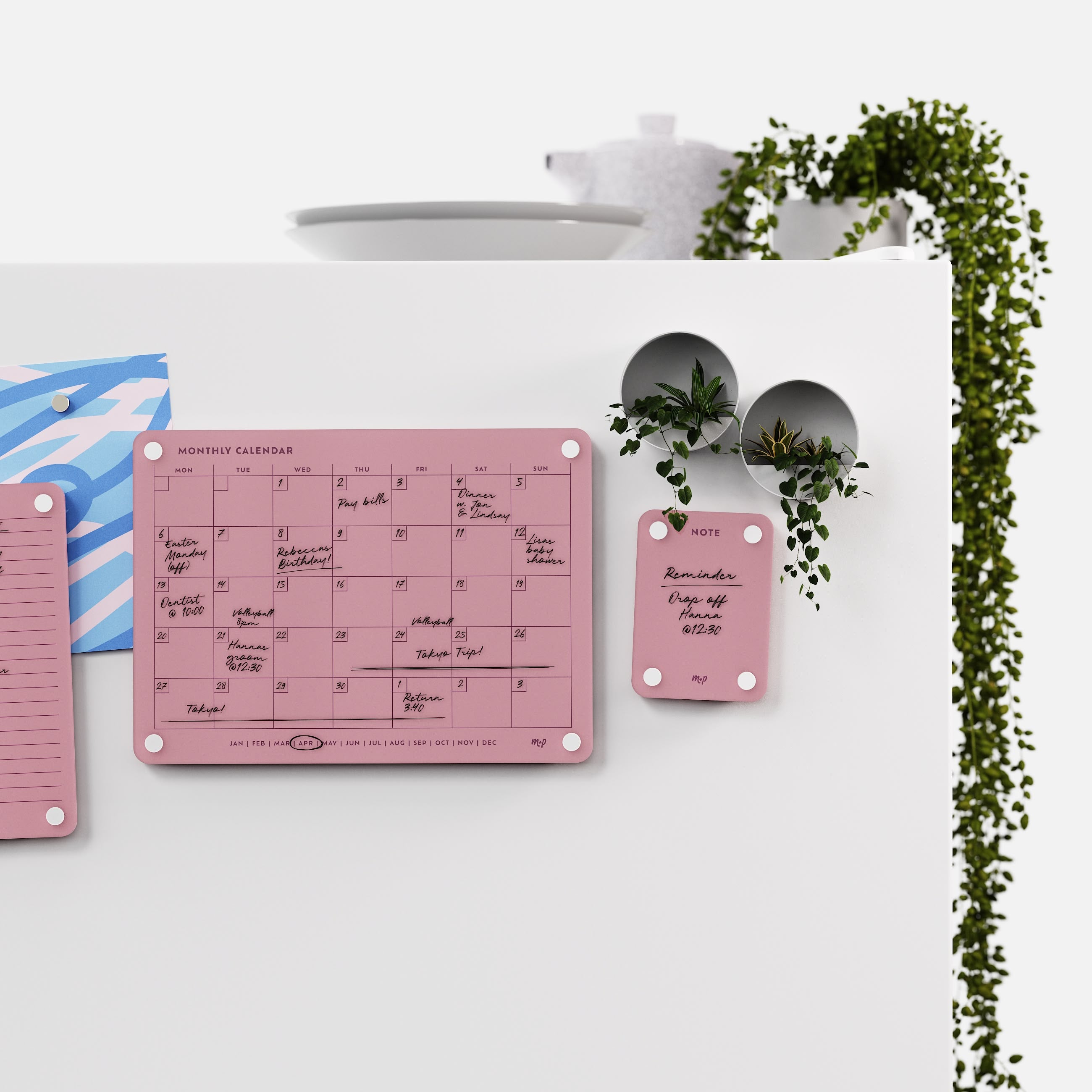 Plum monthly calendar and note board on a white kitchen wall with magnetic planters and trailing ivy